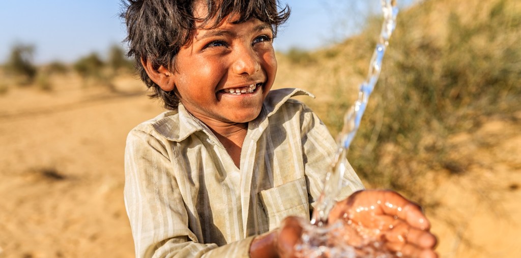 A young child smiling while drinking water from his hands