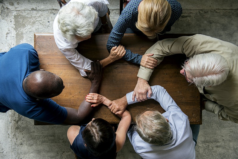 A group of people from all background and ages sitting in a table grabbing each other's hands to symbolize diversity and inclusion