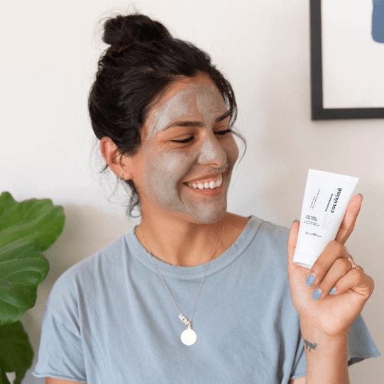 A woman using Cocokind's sea kale clay face mask while holding the product on her hands, smiling 